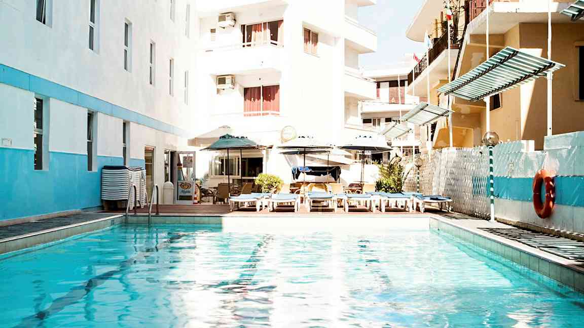 Et utendørsbasseng omgitt av hotellbygninger og solsenger. (AI-generert tekst)