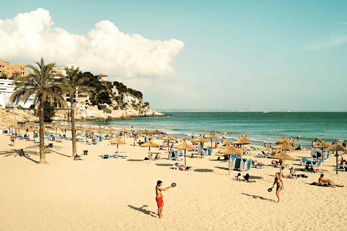 En sandstrand med parasoller, solsenger og personer som nyter solen og havet. Lenger unna ser man en klippe med bygninger. (AI-generert tekst)