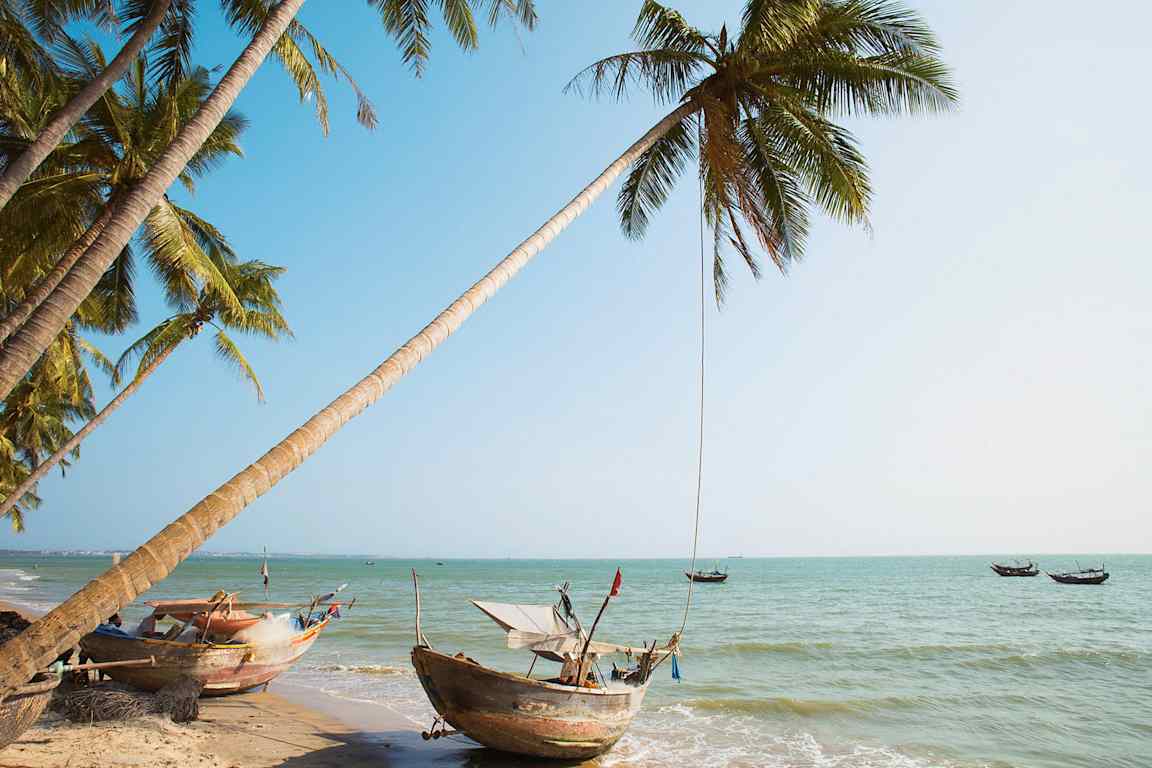 Utsikt over en tropisk strand med palmer og tradisjonelle båter ved vannet. (AI-generert tekst)