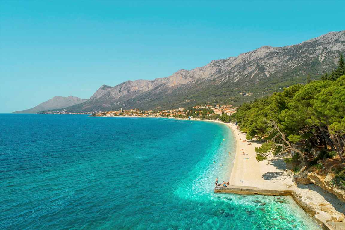 Utsikt over en rullesteinstrand med turkist vann og frodige trær. Fjell og en landsby vises i bakgrunnen. (AI-generert tekst)