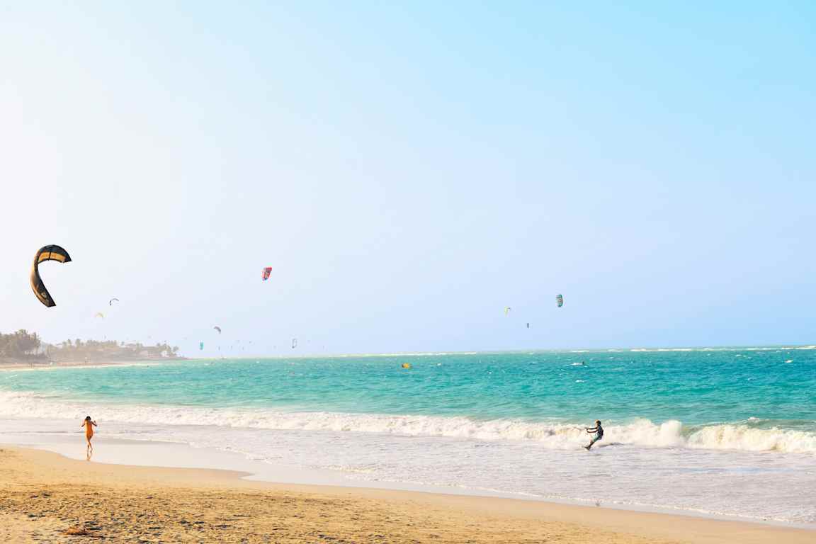 En strand med turkist hav og mange kitesurfere. (AI-generert tekst)