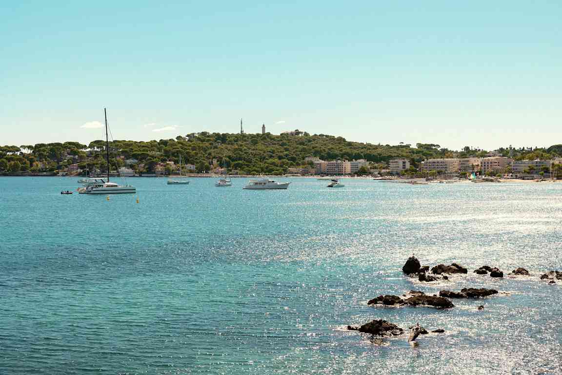 Utsikt over en kystlinje med klart blått vann, flere båter og seilbåter i nærheten av en frodig strand og en by. (AI-generert tekst)
