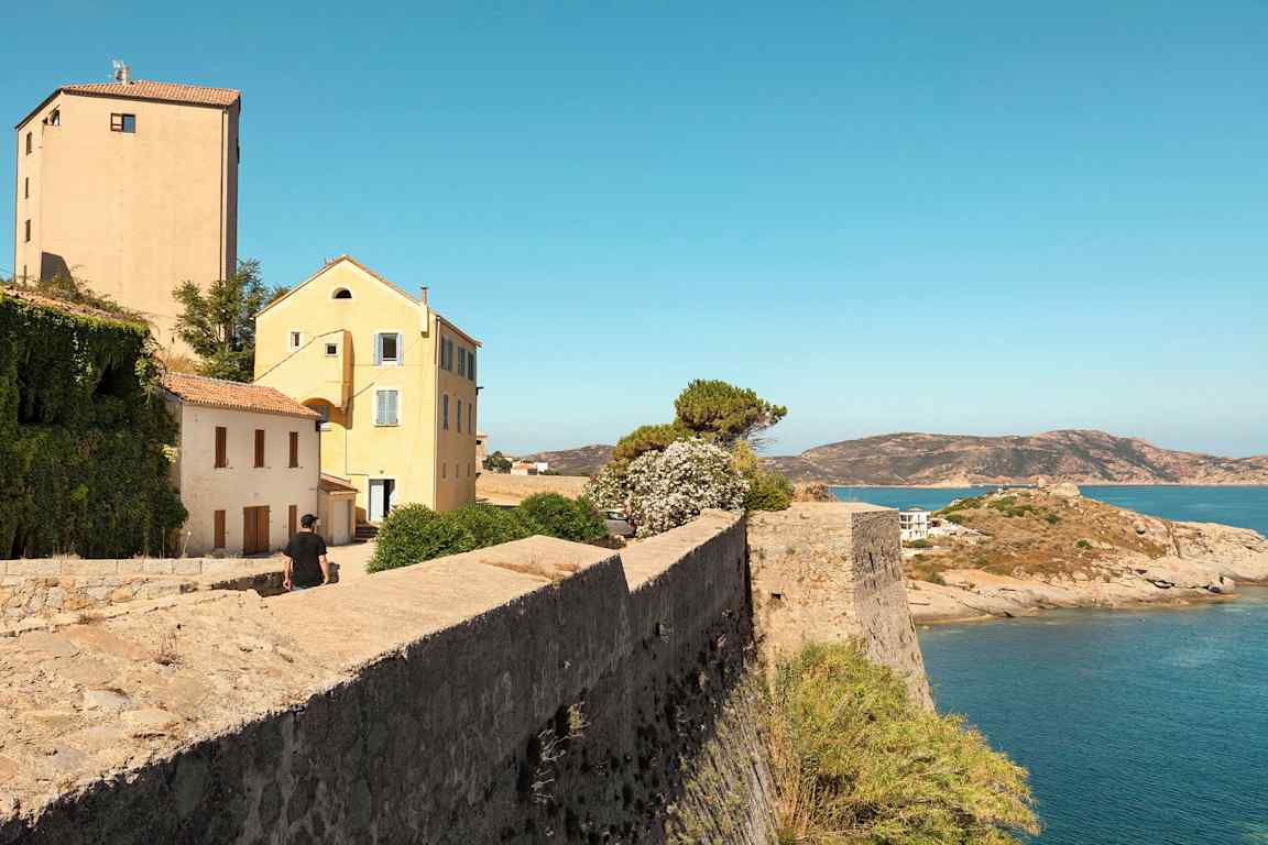 Bilde av en steinmur ved vannet med hus og vegetasjon i bakgrunnen, samt en mann som går mot bygningene. (AI-generert tekst)