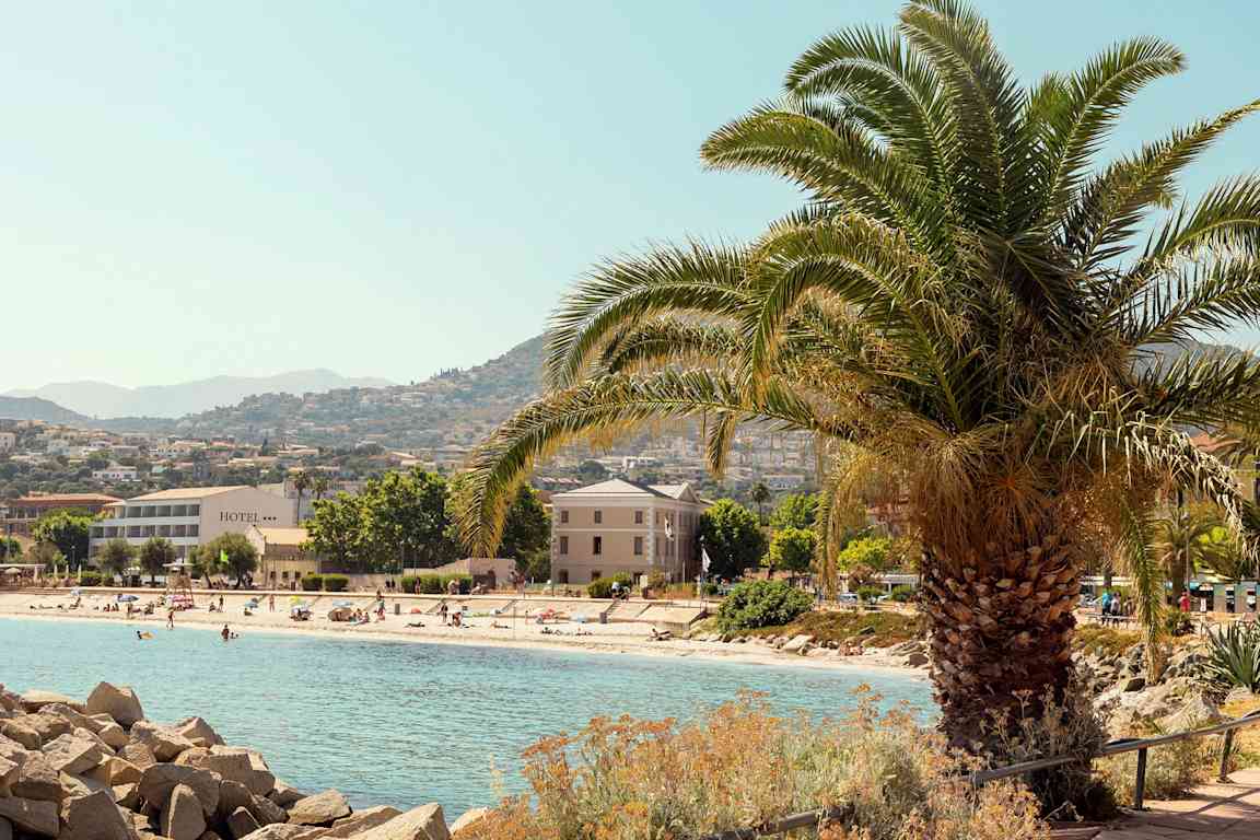 En strand med badegjester, omgitt av hotellbygninger og trær med fjell i bakgrunnen. En stor palme står i forgrunnen. (AI-generert tekst)