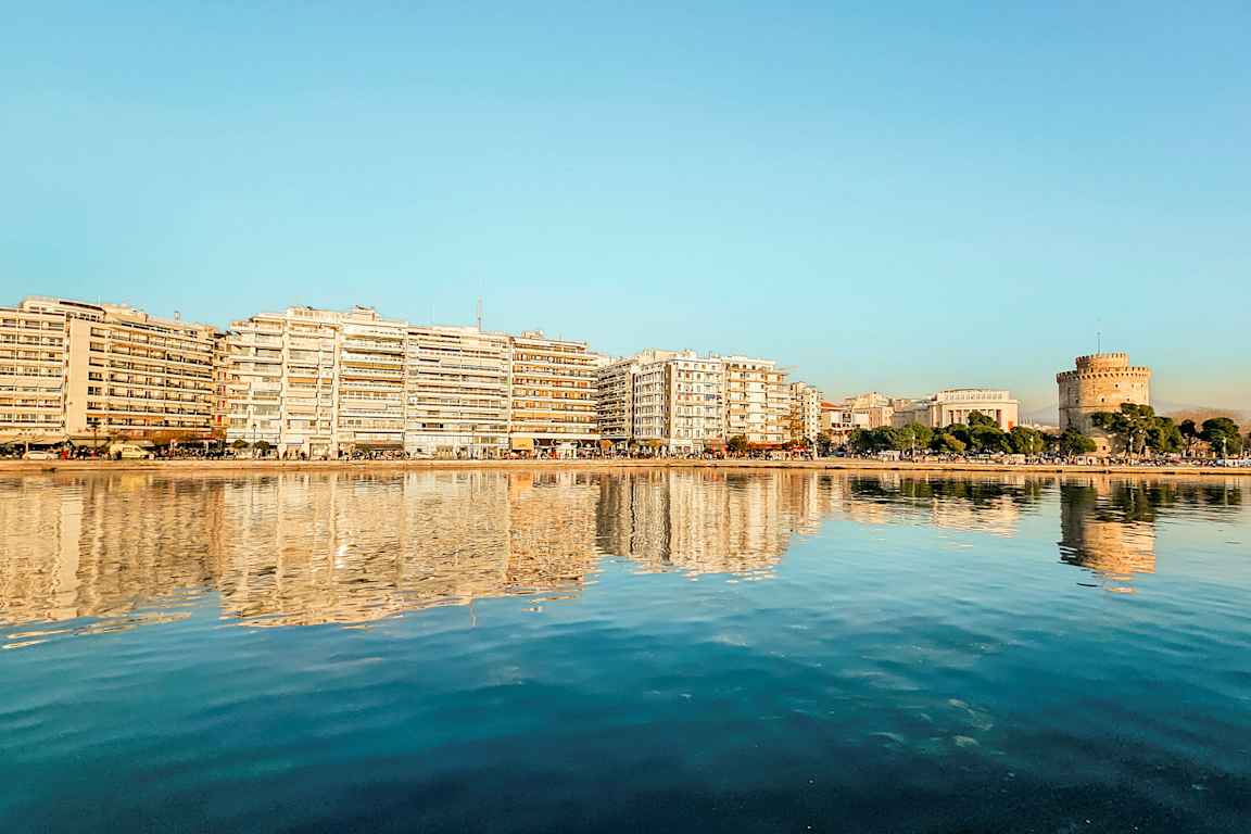 En by ved vannet med bygninger og et rundt tårn reflektert i det rolige vannet. (AI-generert tekst)