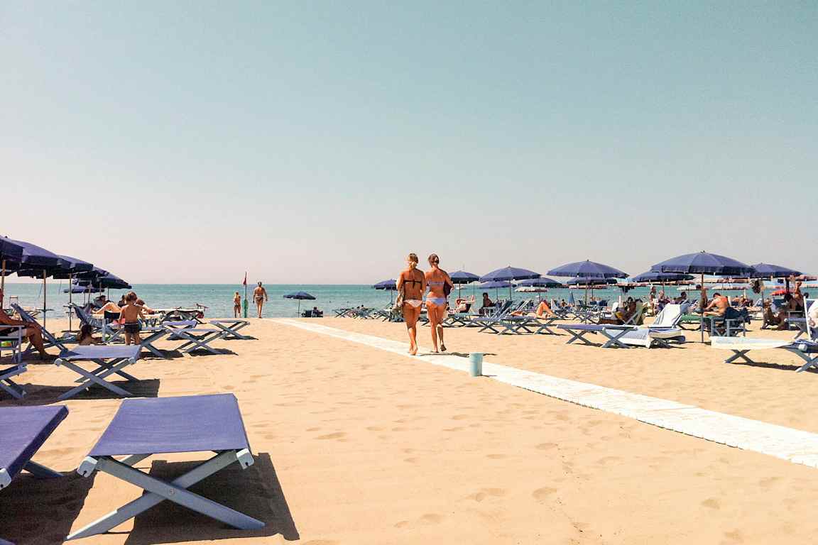 En sandstrand med blå solsenger og parasoller. Folk soler seg og går på stranden med en klar blå himmel i bakgrunnen. (AI-generert tekst)