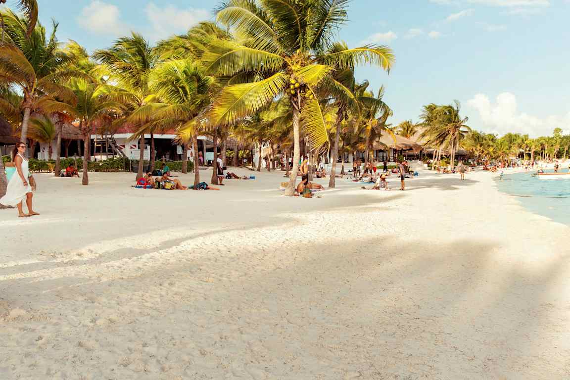 En strand med hvit sand, palmer og mennesker som nyter solen og havet. (AI-generert tekst)