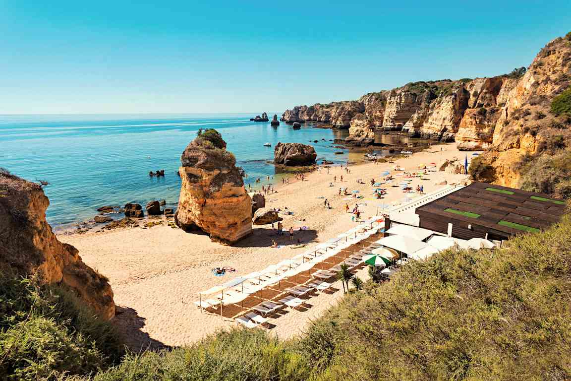 En sandstrand med solsenger og parasoller, omgitt av klippeformasjoner og et stille hav. (AI-generert tekst)