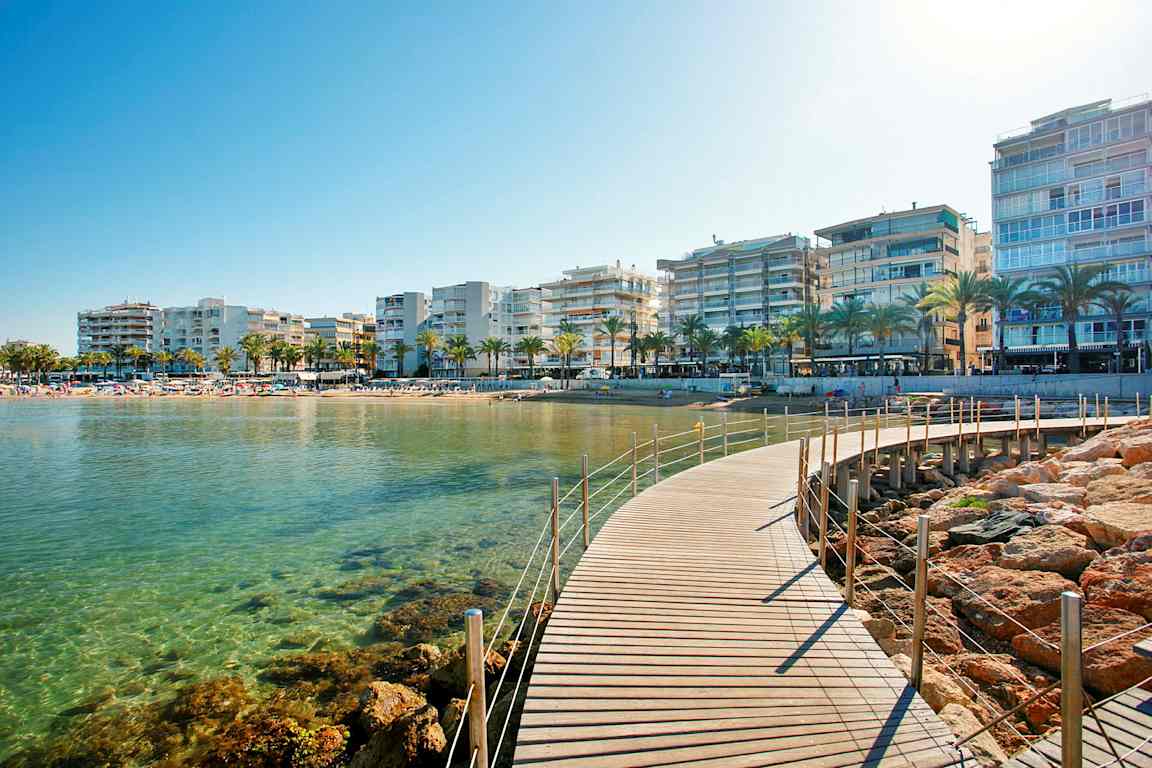 Bilde av en strandpromenade med en gangbro, klart vann, palmer og bygninger i bakgrunnen. (AI-generert tekst)