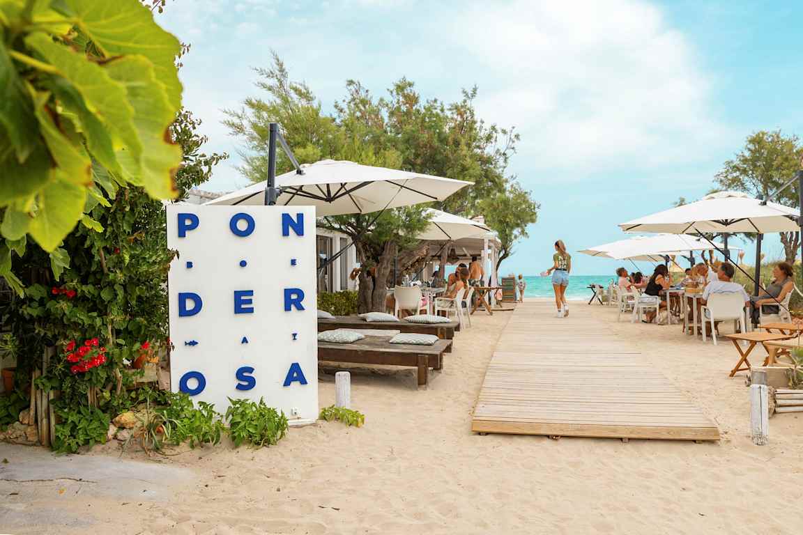 En restaurant ved stranden med bord og parasoller, en tresti fører mot vannet. (AI-generert tekst)