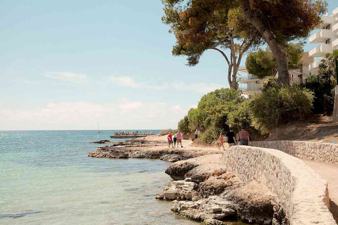 Steinstrand med svingende gangvei nært havet, omgitt av trær og bygninger. (AI-generert tekst)