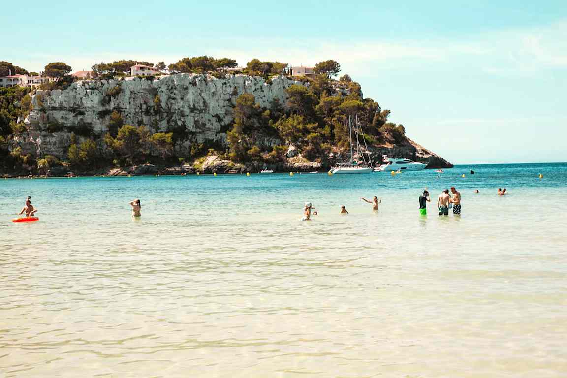 En strand med mennesker i vannet foran en ås med klipper og noen hus samt båter i bakgrunnen. (AI-generert tekst)