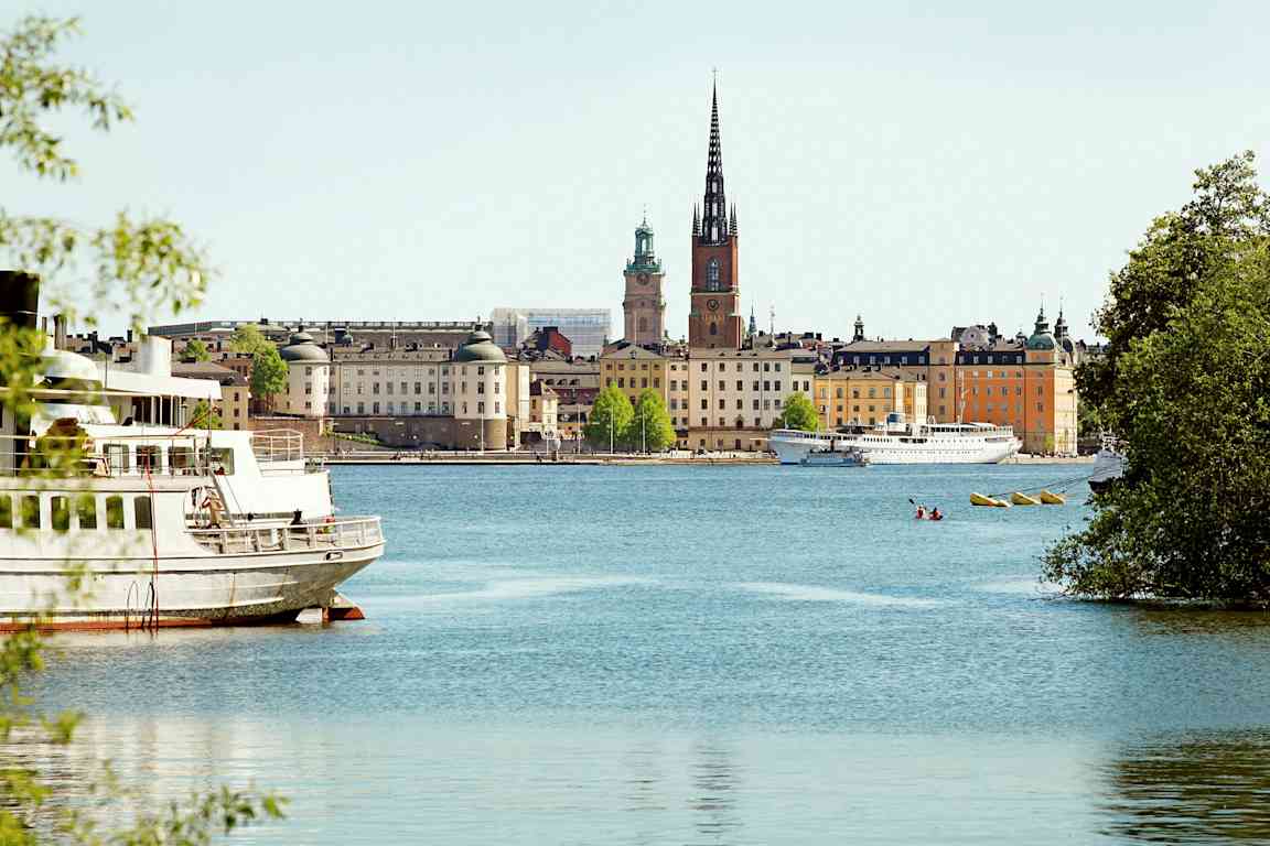 En byutsikt med bygninger, vann og en kirke med en høy spir, sett fra et sted ved vannet. (AI-generert tekst)