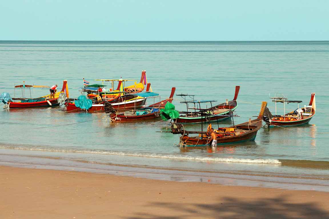 Flere fargerike båter ligger fortøyd på vannet nær en sandstrand. (AI-generert tekst)