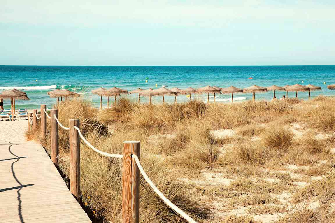 Strand med solsenger og parasoller med stråtak ved havet. En tresti fører mot stranden gjennom sanddyner. (AI-generert tekst)