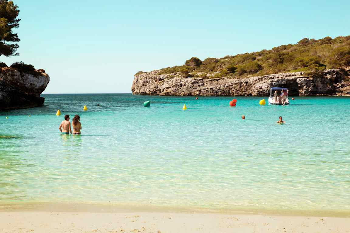 En sandstrand med mennesker i det rolige, turkise vannet. I bakgrunnen er en båt og klipper dekket av grønt. (AI-generert tekst)
