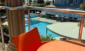 Utsikt fra en balkong med et oransje bord og stol i forgrunnen, et basseng og et hotell i bakgrunnen. (AI-generert tekst)