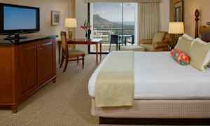 Et hotellrom med en stor seng, en TV, et skrivebord og en stol. I bakgrunnen sees en balkong med utsikt over fjell og hav. (AI-generert tekst)