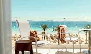 Utsikt fra en balkong mot en strand med solsenger og parasoller, palmer samt hav og fjell i det fjerne. (AI-generert tekst)
