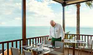 En spisesal med dekkede bord på en terrasse med utsikt over havet. (AI-generert tekst)