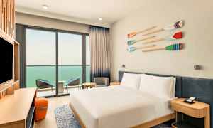 Et hotellrom med en hvitredd dobbeltseng, en TV på en benk og en sittegruppe ved en balkong med utsikt over havet. (AI-generert tekst)