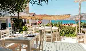 En utendørs servering med hvite trestoler og bord, dekorert med blomster, og en utsikt over havet. (AI-generert tekst)