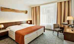 Et hotellrom med en dobbeltseng som har et oransje sengeteppe og to hvite puter. Det er nattbord med lamper på begge sider og et stort vindu med gardiner i bakgrunnen. Et skrivebord med en stol og diverse gjenstander står til høyre. (AI-generert tekst)