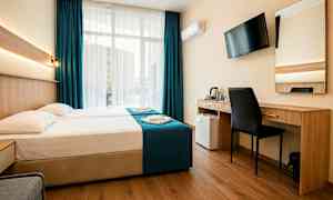 Et hotellrom med en dobbeltseng, skrivebord, stol, veggmontert TV og store vinduer med mørkeblå gardiner. (AI-generert tekst)