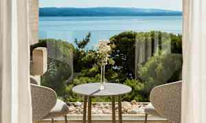 En borddekorasjon med blomster ved et bord foran vinduer med utsikt over havet og vegetasjon. (AI-generert tekst)