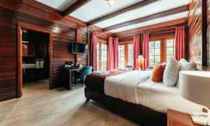 Hotellrom med treinteriør, en bred seng med hvite og fargerike puter, røde gardiner og vinduer med utsikt. (AI-generert tekst)