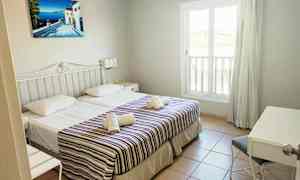 Et hotellrom med to enkeltsenger, dekorerte med stripete overkast og håndklær. Et vindu og en skrivebordshjørne er synlig. (AI-generert tekst)