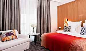 Hotellrom med seng, mønstret pute, rødt teppe, stol og bord med blomster. (AI-generert tekst)