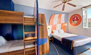 Et hotellrom med en tre køyeseng, en dobbeltseng, dekor i blått og oransje og et veggmaleri med sjørøvermotiv. (AI-generert tekst)