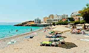 En solrik strand med parasoller, folk som bader og bygninger ved strandkanten. (AI-generert tekst)