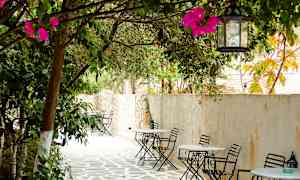 Et skyggefullt uteområde med bord og stoler av jern plassert langs en steinbelagt gangsti under tregrener dekorert med rosa blomster. (AI-generert tekst)