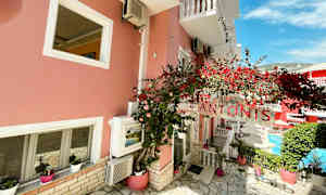 En rosa bygning med hvite vinduer, skilt med teksten 'Antonis' og røde blomster. (AI-generert tekst)