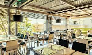 En restaurant med flere dekkede bord og stoler, med vinduer mot en byutsikt. (AI-generert tekst)