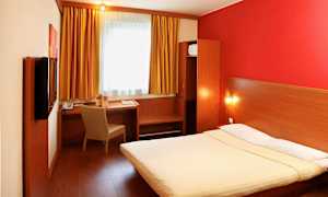 Et hotellrom med en dobbeltseng, skrivebord og stol samt et skap. Vinduet har gule gardiner. (AI-generert tekst)