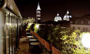 En takterrasse med bord og stoler omgitt av grønne planter, med kirketårn og kupler som vises i bakgrunnen om kvelden. (AI-generert tekst)