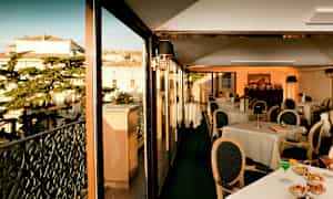 En restaurant med glassvegger og utsikt over bygninger og trær. (AI-generert tekst)