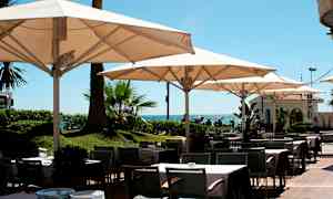 Utendørs restaurantområde med bord, stoler og parasoller nær havet. (AI-generert tekst)