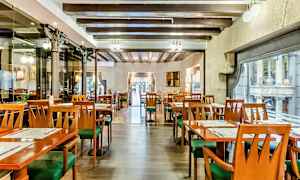Et interiør av en restaurant med tomme bord og stoler, trebjelker i taket og store vinduer mot en gate. (AI-generert tekst)