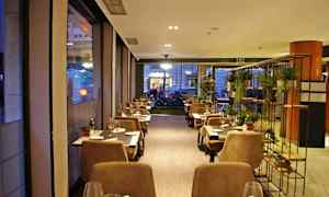 En koselig restaurant med bord og stoler arrangert langs vinduer og et dekorativt stativ med planter. (AI-generert tekst)