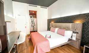 Et lyst hotellrom med en dobbeltseng oppredd med hvitt sengetøy og rosa sengeteppe, et skrivebord med en stol og en dekorativ vegg bak sengen. (AI-generert tekst)
