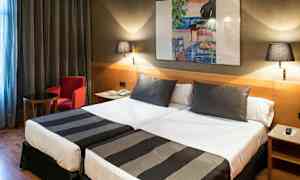 Et hotellrom med to enkeltsenger, begge oppredd med svart-hvite sengetepper. På veggen henger et fargerikt maleri, og rommet har også et lite bord, en rød lenestol og to vegglamper. (AI-generert tekst)