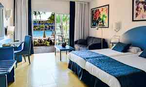 Et hotellrom med en stor seng, sittegruppe og utsikt over et basseng gjennom glassdører. (AI-generert tekst)