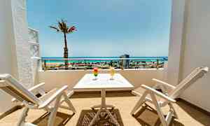 Terrasse med to hvite stoler og et bord, to drinker og utsikt over stranden og havet. (AI-generert tekst)