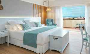 Et lyst hotellrom med en stor dobbeltseng, stol og et bord, samt en balkong med utsikt over havet. (AI-generert tekst)