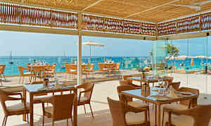 Bilde av en restaurant med flettede stoler og glassbord, beliggende ved havet med utsikt mot en blå vannhorisont. (AI-generert tekst)