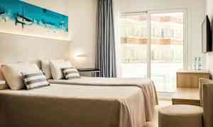 Et hotellrom med to enkeltsenger, hvite vegger og et vindu som vender mot en balkong. Et maleri pryder veggen over sengene. (AI-generert tekst)